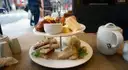 A tiered stand with sandwiches and scones next to a white teapot