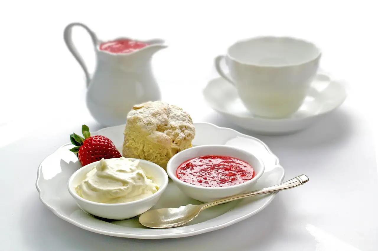 A scone with cream, jam and a strawberry on a plate beside a teacup