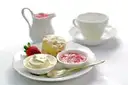 A scone with cream, jam and a strawberry on a plate beside a teacup