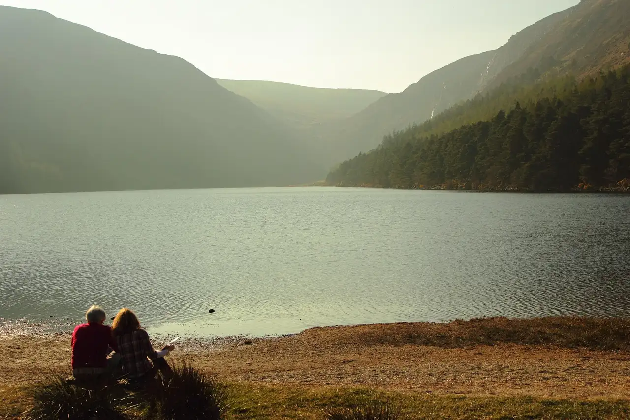 Two people sitting on the shore of the Upper Lake at Glendalough, with wooded hills behind