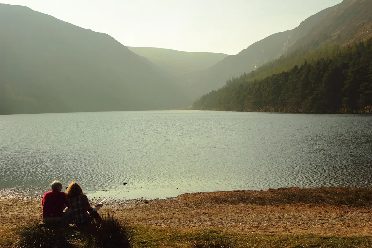 Two people sitting on the shore of the Upper Lake at Glendalough, with wooded hills behind