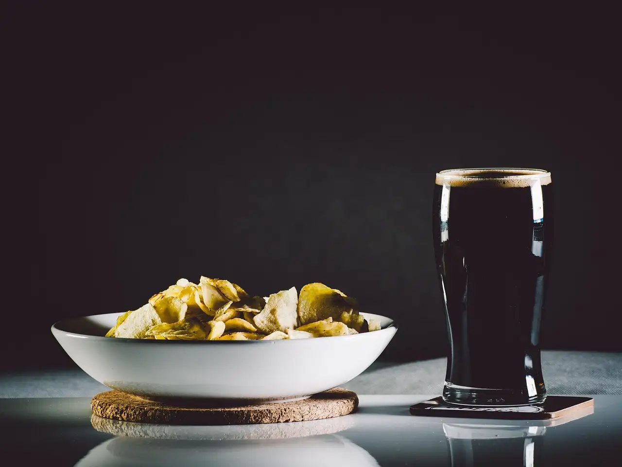A pint of dark stout with a creamy head on a table, next to a bowl of crisps