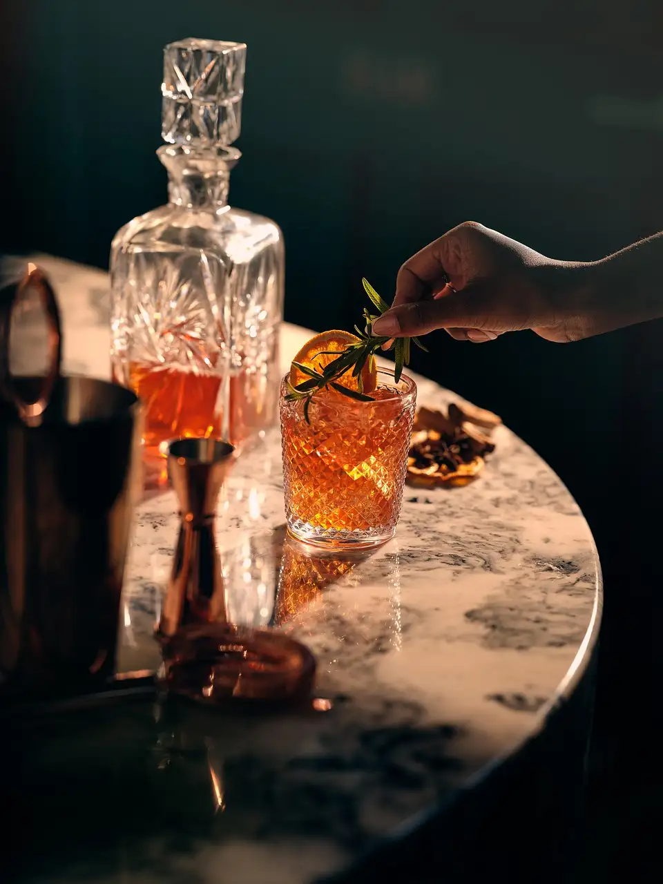 A bartender adds a garnish to an amber cocktail next to a crystal decanter