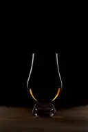 A tulip-shaped whiskey glass with a little amber whiskey, lit against a black background