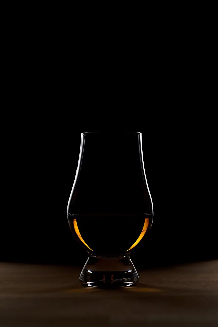 A tulip-shaped whiskey glass with a little amber whiskey, lit against a black background