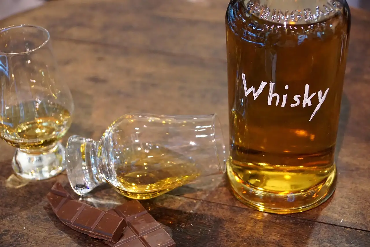 A bottle of whisky, two tasting glasses and squares of chocolate on a wooden table