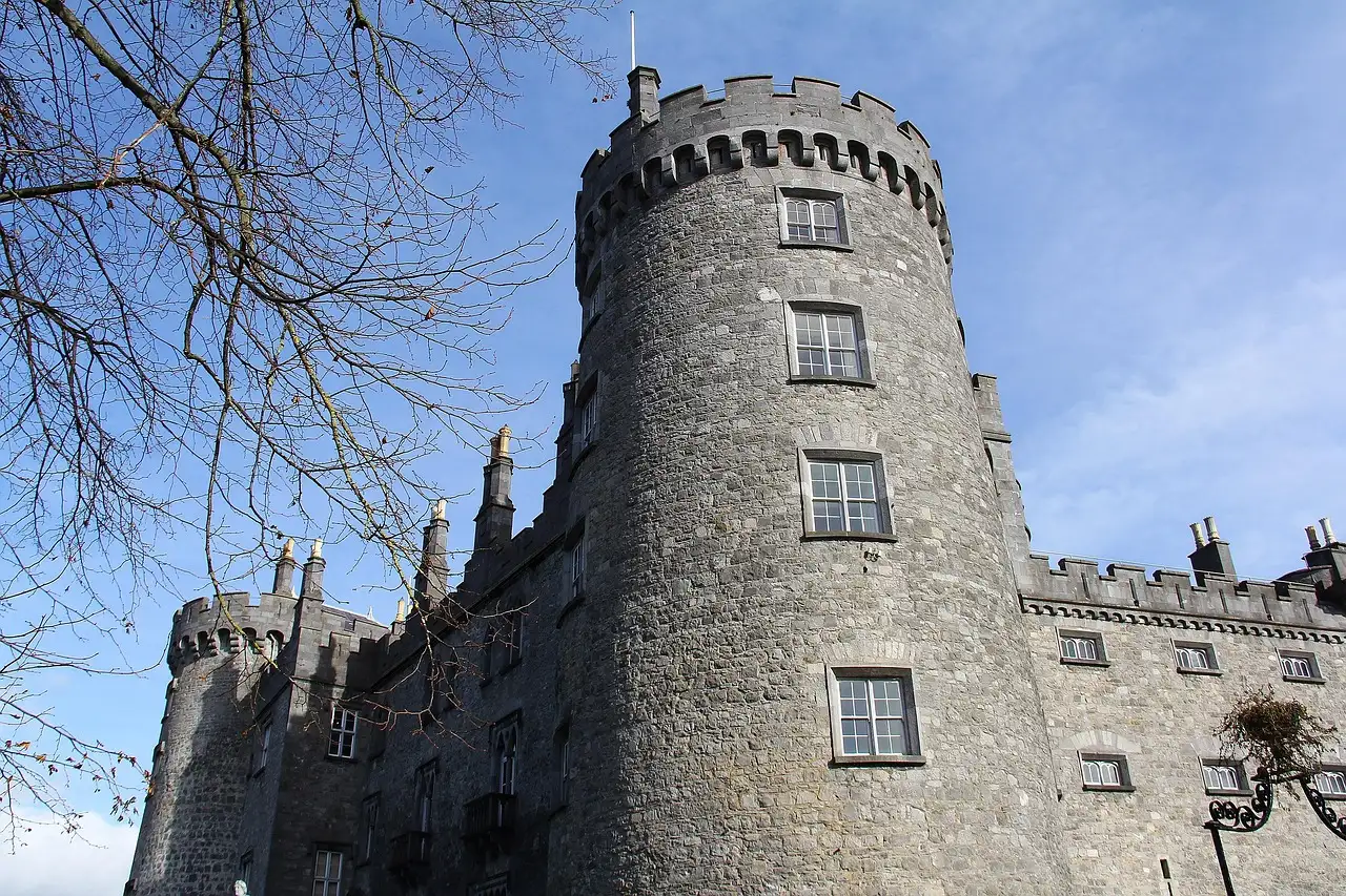 A round stone tower of Kilkenny Castle against a blue sky, with bare branches in front