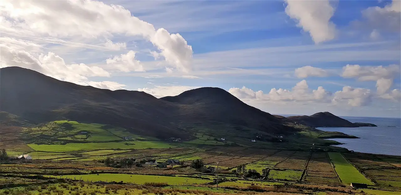 Green fields and dark hills by the sea on the Ring of Kerry under a cloudy blue sky