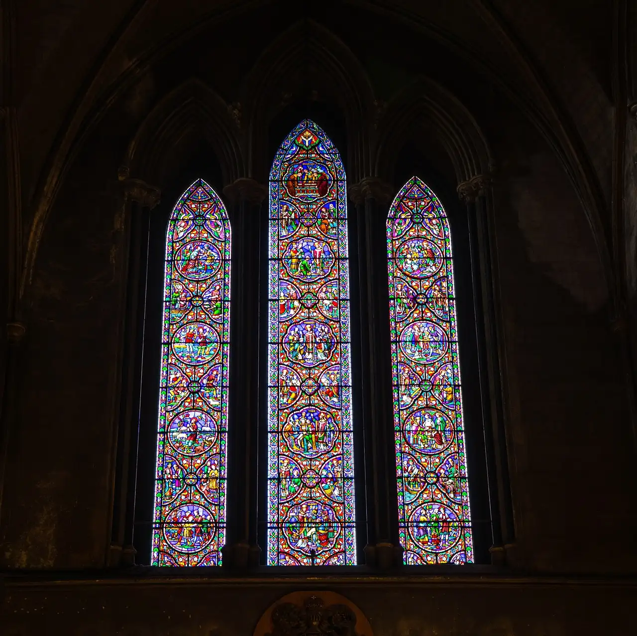 Three tall stained glass windows glowing in a dark stone wall