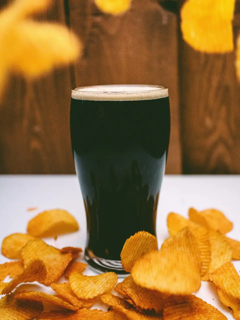 A pint of dark stout with a creamy head on a table, next to a pile of crisps
