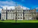 A grey stone Trinity College building behind a wide green lawn and a young tree